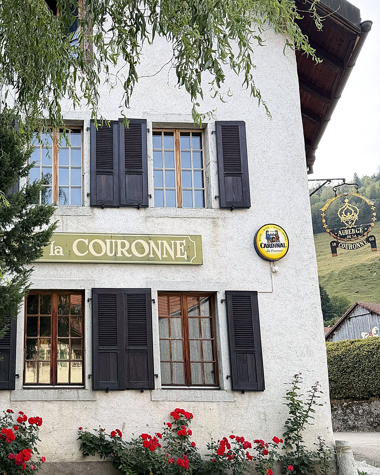 Façade blanche de l'Auberge de la Couronne aux volets bruns, avec son enseigne en fer forgé et des rosiers rouges